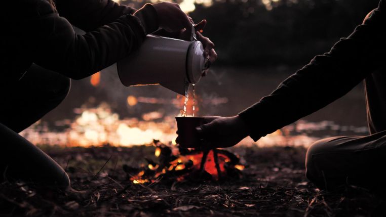 People pouring a warm drink around a campfire